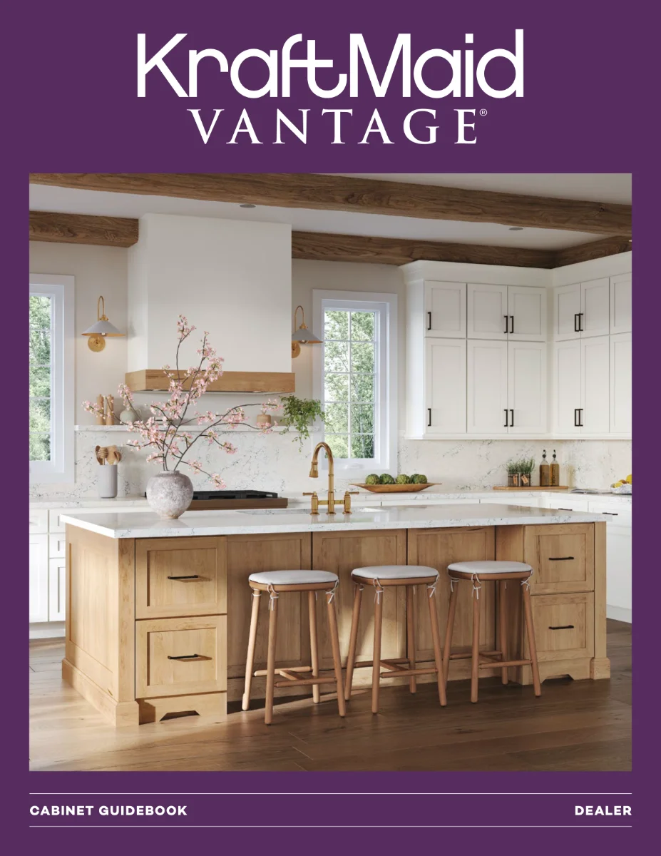 The cover of a KraftMaid Vantage brochure with a large, well-lit kitchen with white shaker cabinets, a wooden island, and three modern bar stools. The room includes exposed wood ceiling beams and brass fixtures.